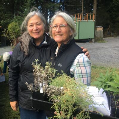 two women in black vests holding green plants from Salish Seeds Project plant sale